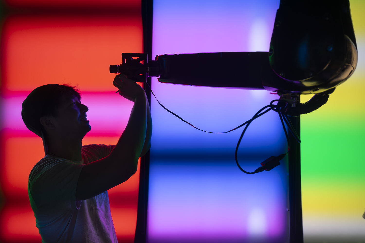 Student is adjusting a robot arm hanging from the ceiling of the lab. The background is covered in large light panels, lit in different colors.