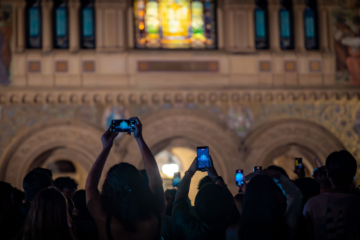Students in silhouette hold up their phone cameras to shoot the illuminated stained glass of Memorial Church.