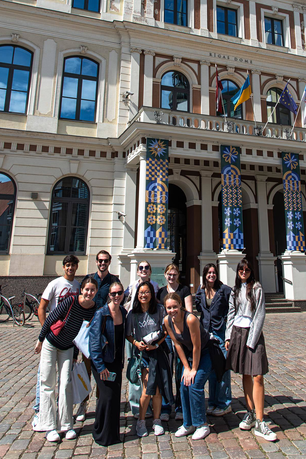 A group of eight people poses for a photo in front of the Rigas Dome building, decorated with vibrant banners.