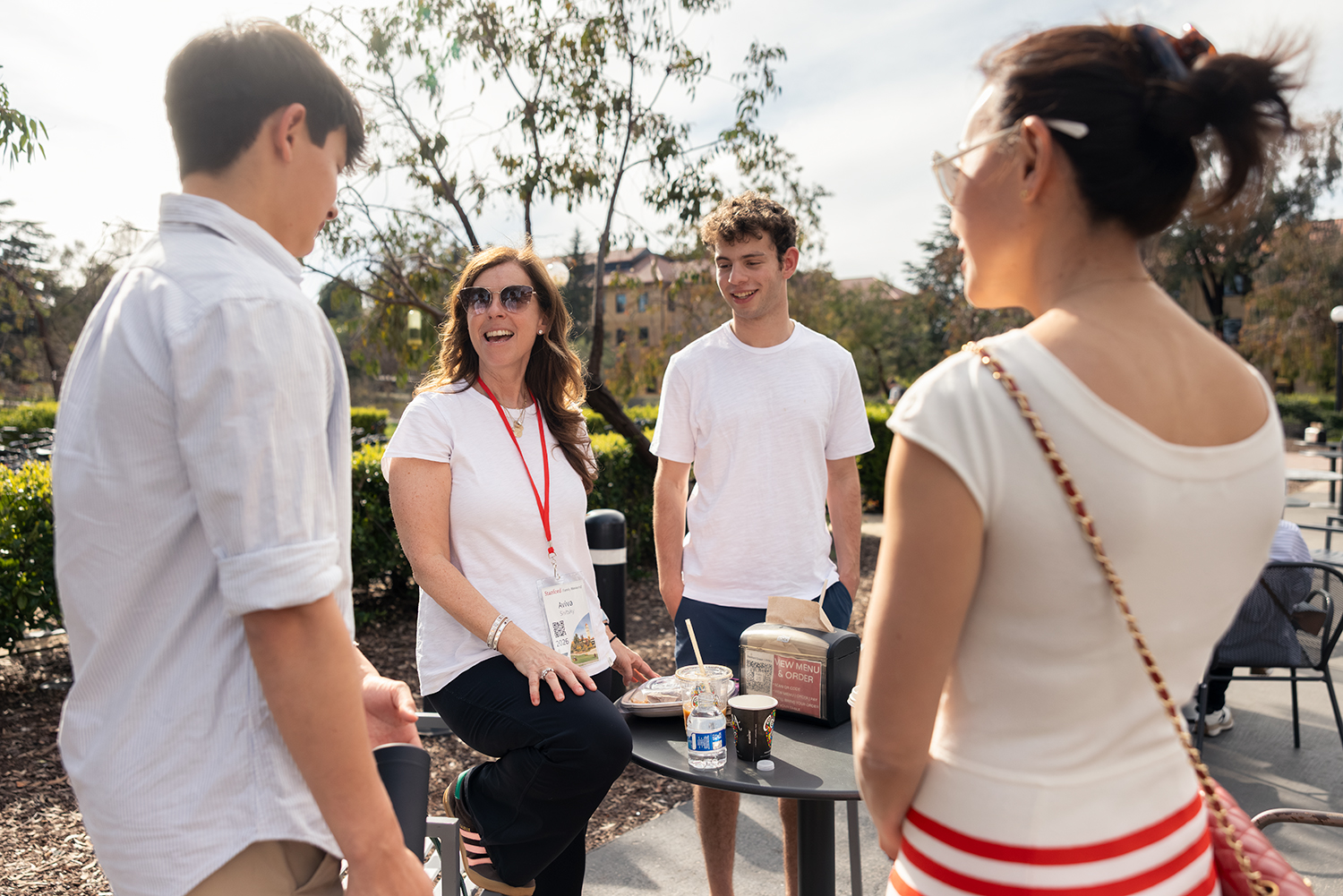 A group of four people engage in conversation outdoors at a table, with snacks and drinks visible.