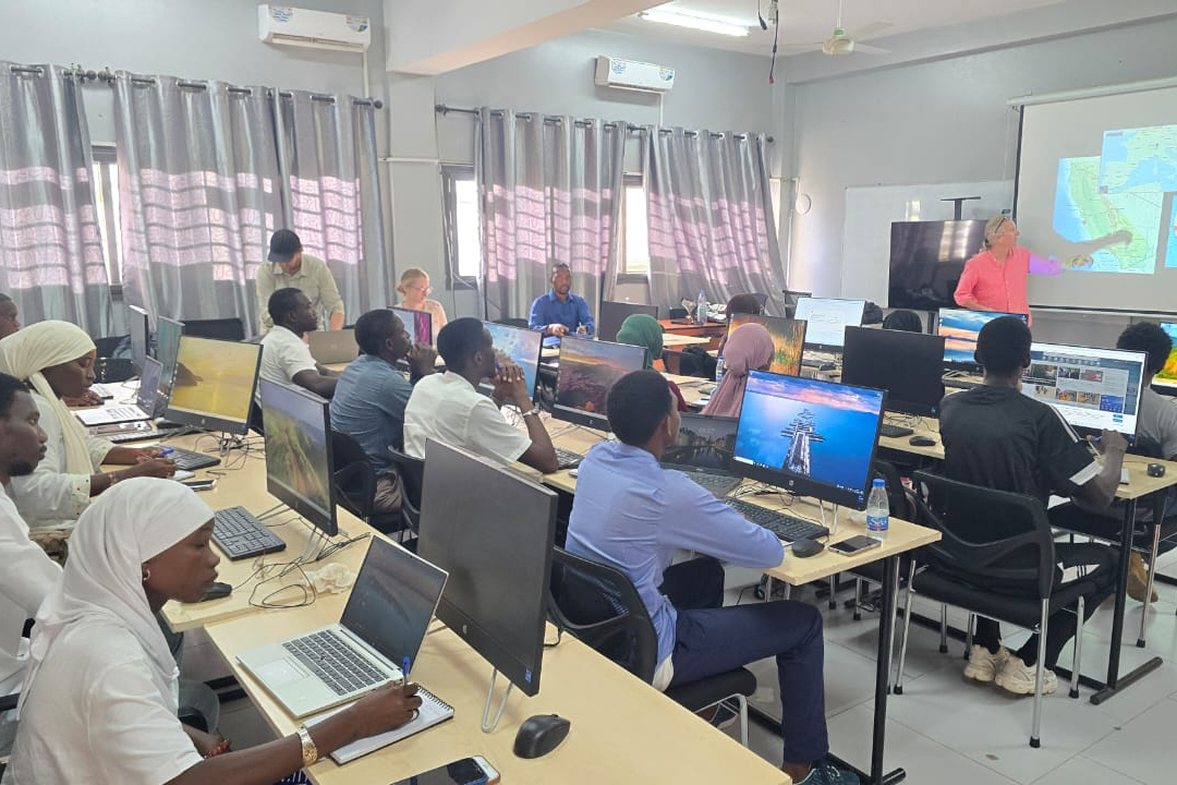 Classroom filled with students at computers, listening to instructor in front of a screen giving a presentation.