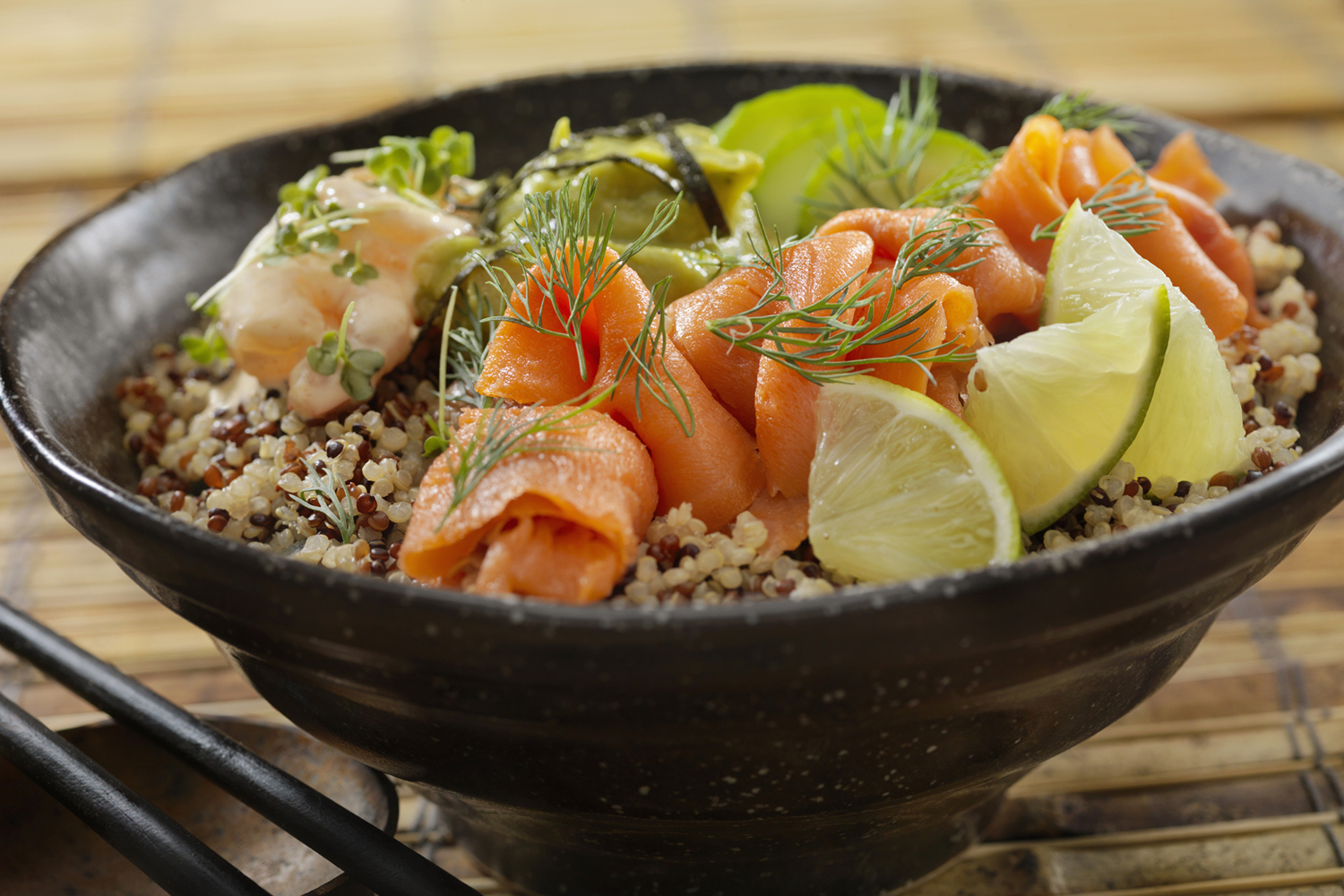 A black bowl filled with quinoa, topped with smoked salmon, green herbs, and lime wedges.