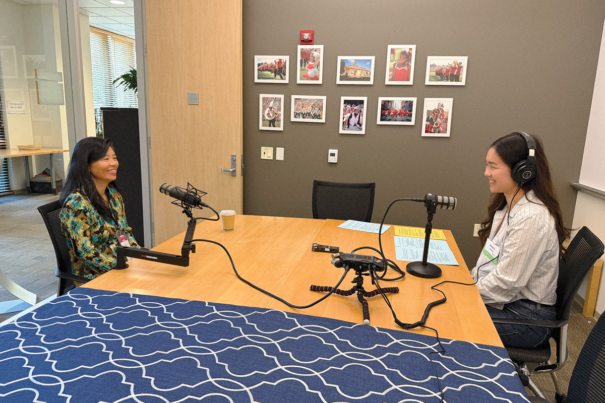 Two women sit across from each other at a table with microphones, recording a podcast in an office setting.