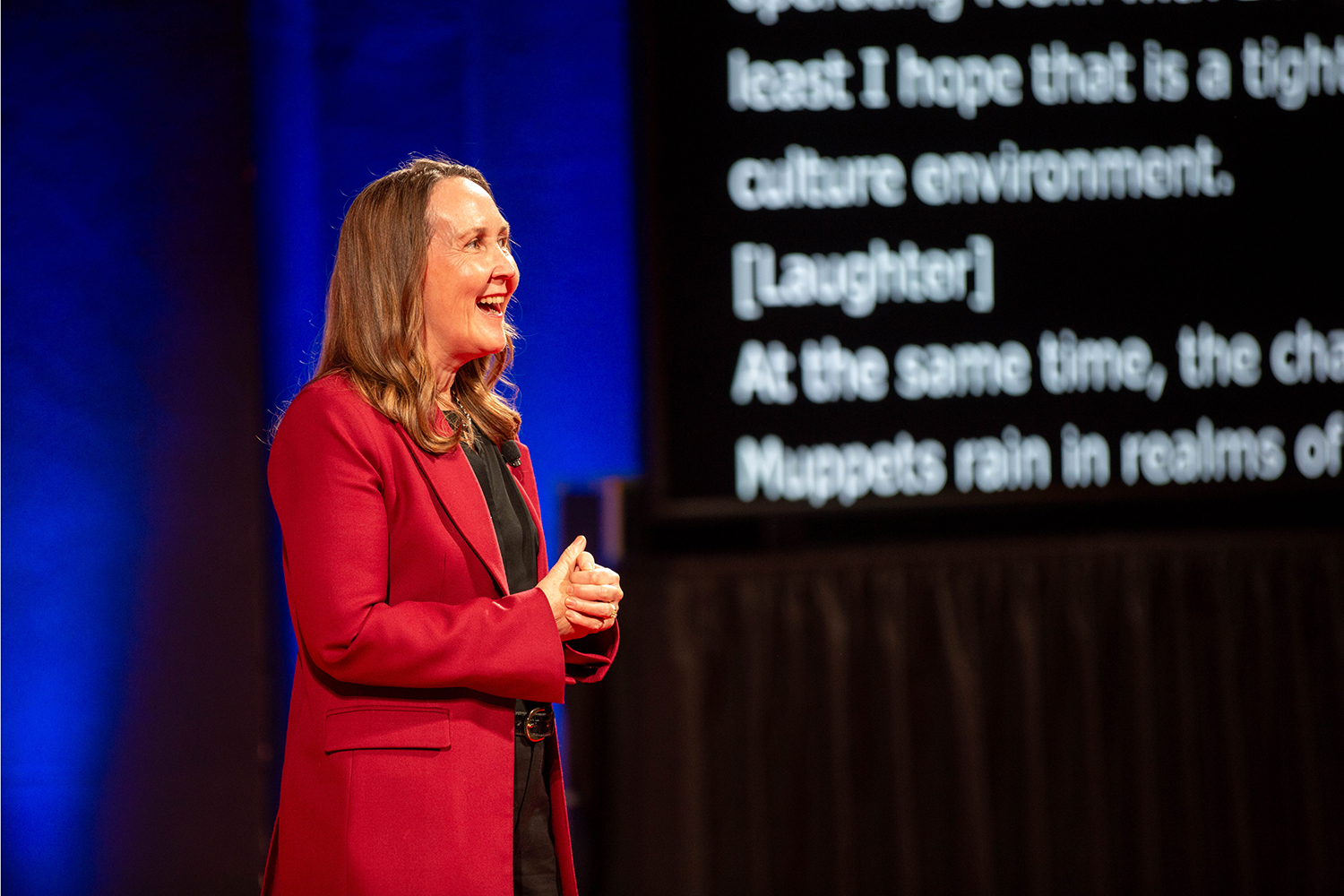 Provost Jenny Martinez in red blazer speaking on stage with projected text visible behind her.