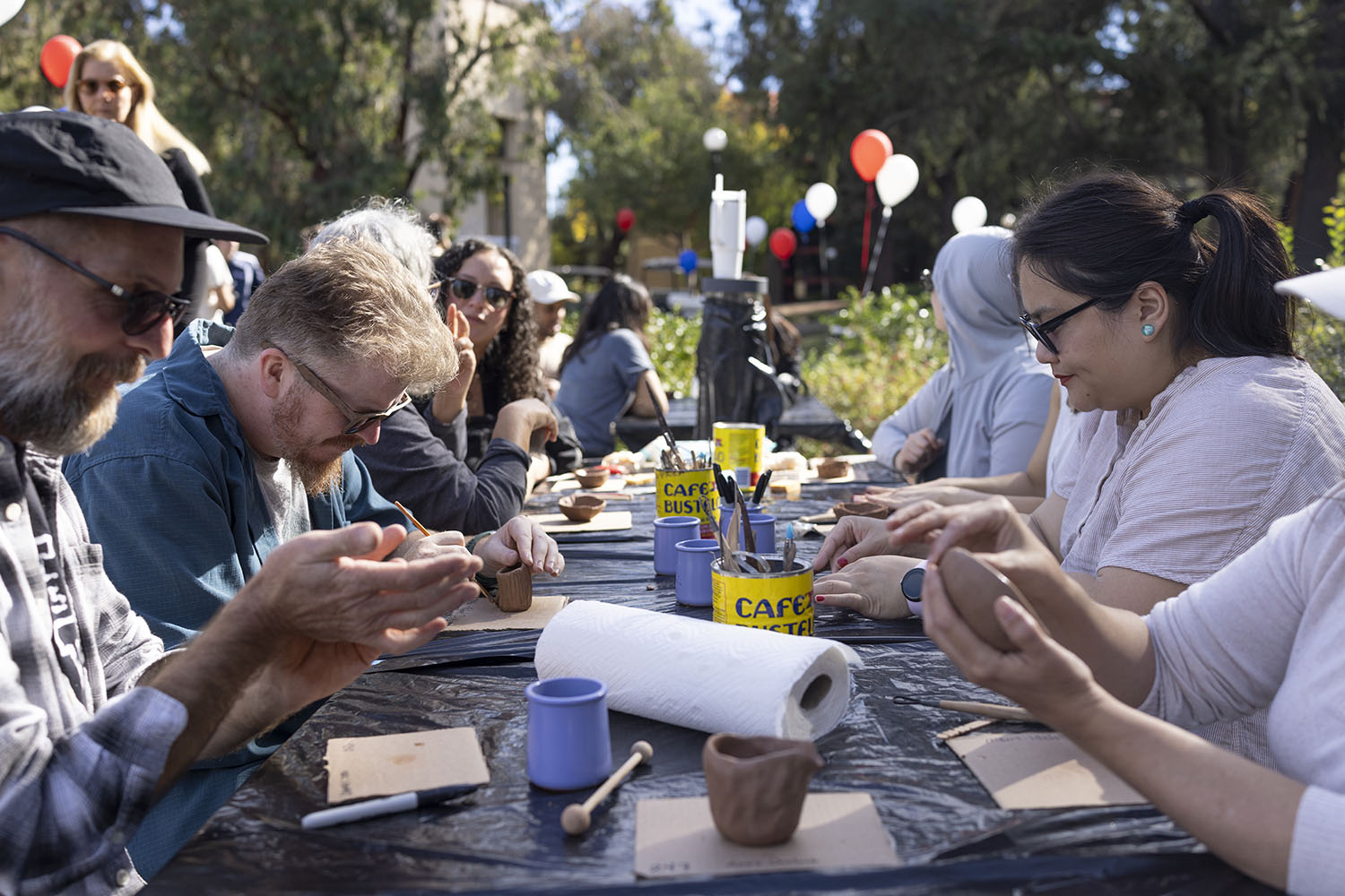 People work on pottery projects together at an outdoor community event with colorful balloons in the background.