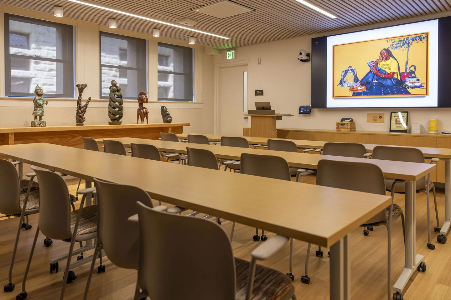 Modern classroom with wooden tables, rolling chairs, African sculptures on display, and a projected artwork on the front screen.