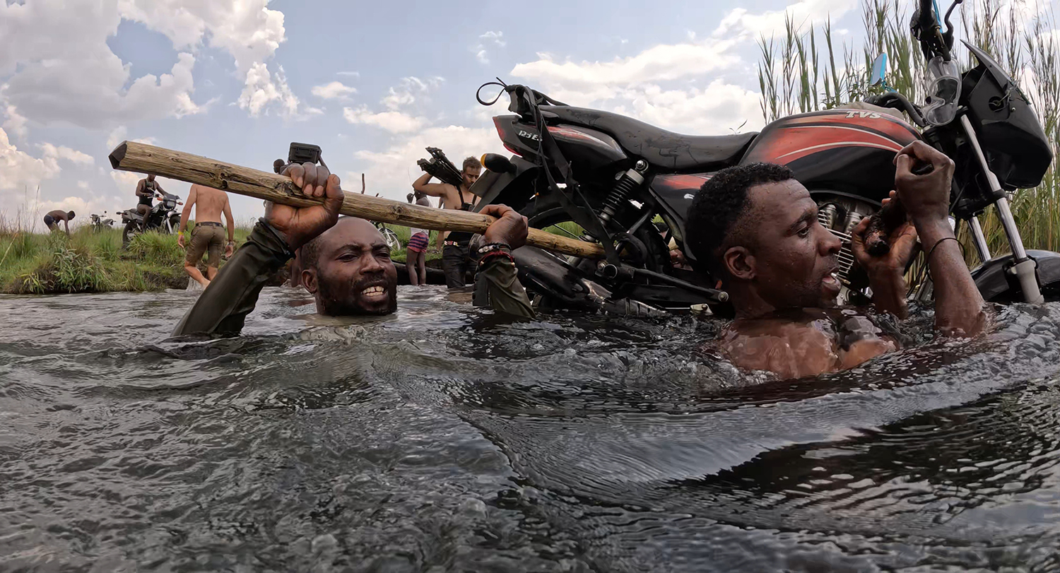 Men wade through deep water, helping to lift a motorcycle with a wooden pole.