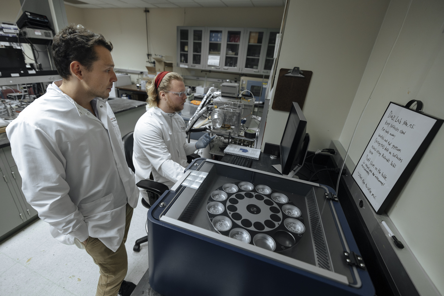 Two scientists in lab coats analyze data near a machine with multiple canisters in a research laboratory.