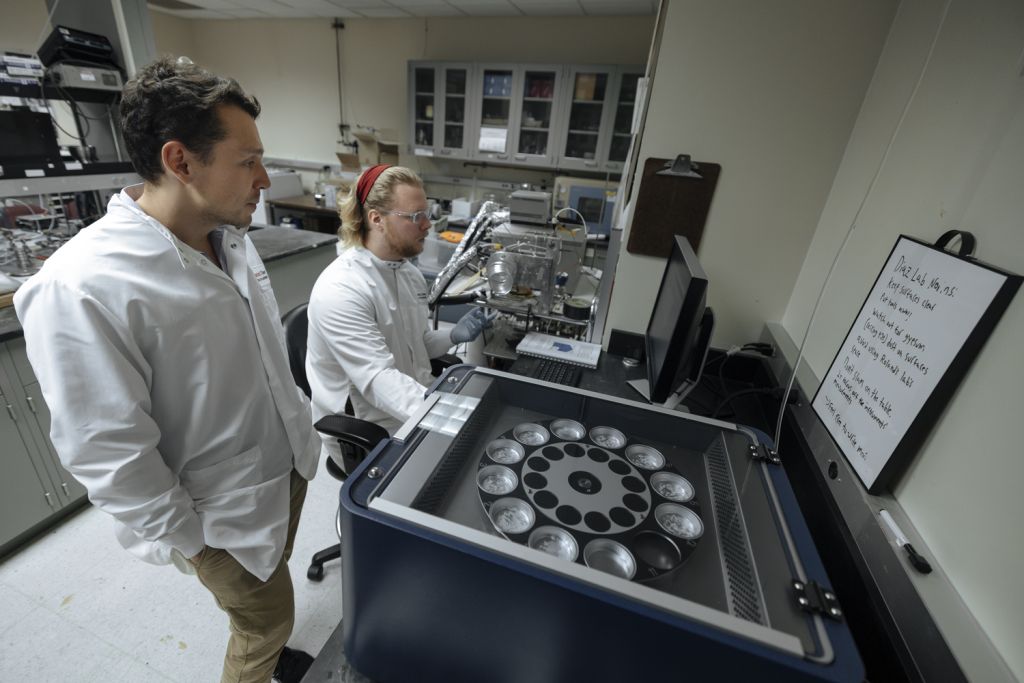 Two scientists in lab coats analyze data near a machine with multiple canisters in a research laboratory.
