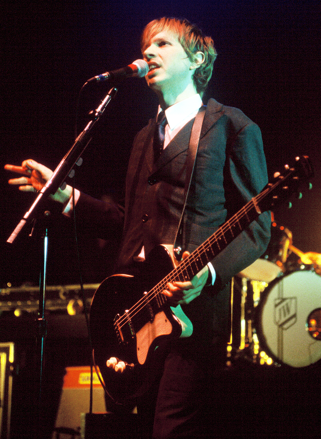 Beck sings into a microphone while holding an electric guitar on stage, performing for an audience.