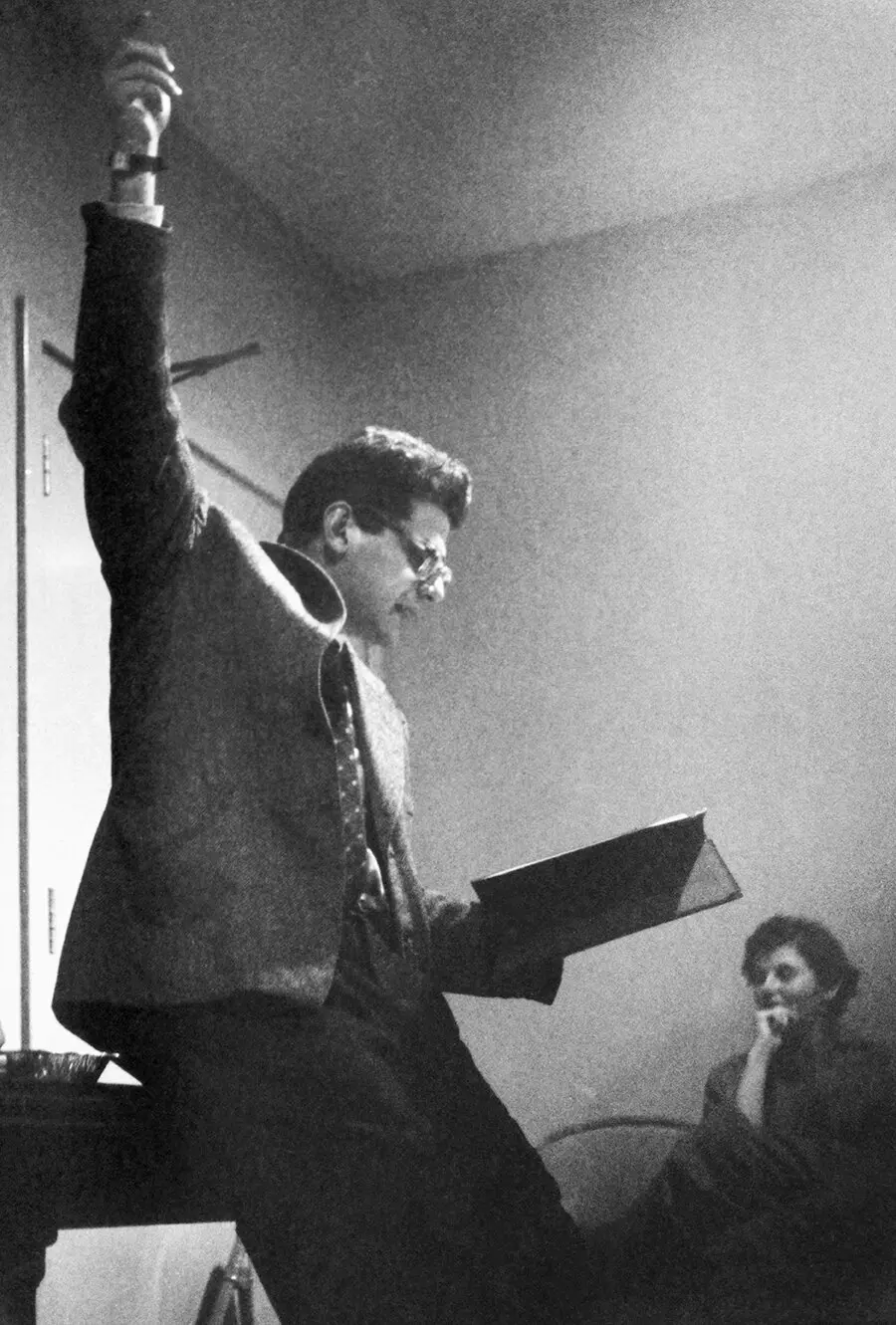 Young Allen Ginsberg with one arm raised and holding a book, passionately reading his poetry to an audience seated in a dimly lit room.
