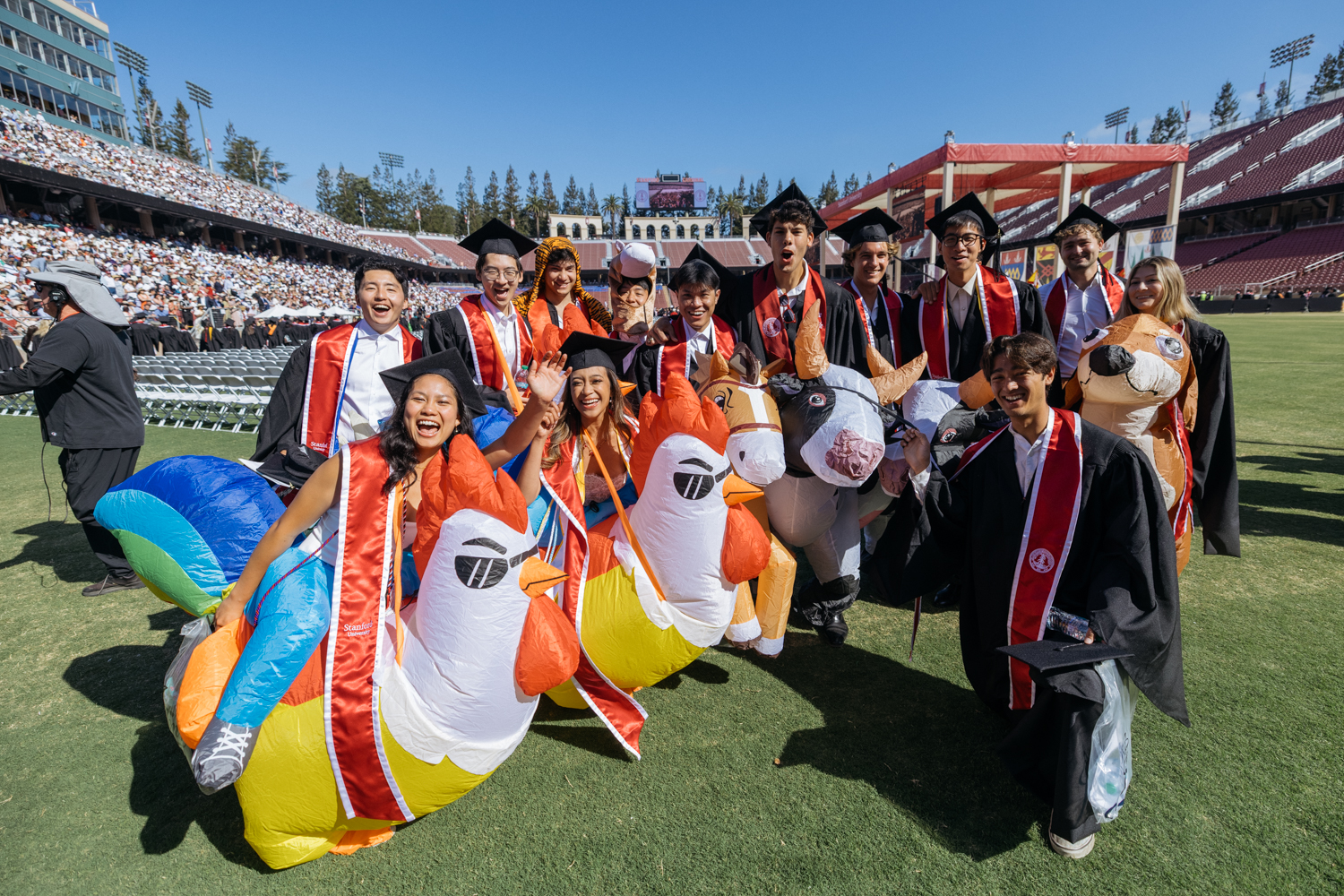 Open video Graduates pose with inflatable animal costumes, celebrating at a graduation ceremony on a sunny day in a stadium. in a modal