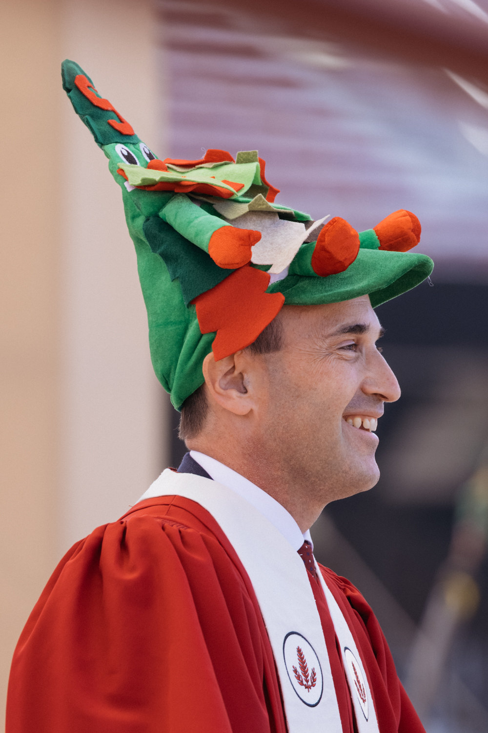 Smiling President Levin in red academic robe wears festive green and red hat shaped like Stanford Tree mascot.