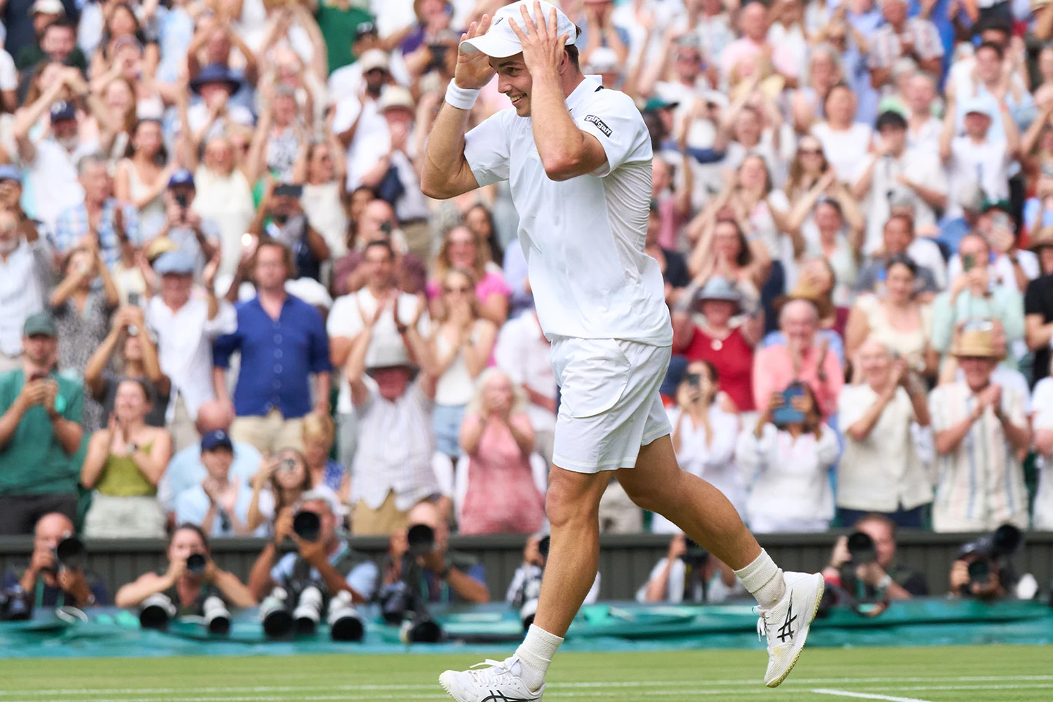 Arthur Fery smiles and holds his cap on his head as a packed Wimbledon crowd stands and applauds behind him.