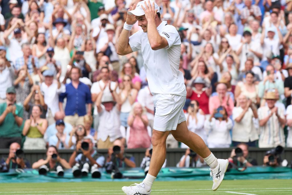 Arthur Fery smiles and holds his cap on his head as a packed Wimbledon crowd stands and applauds behind him.