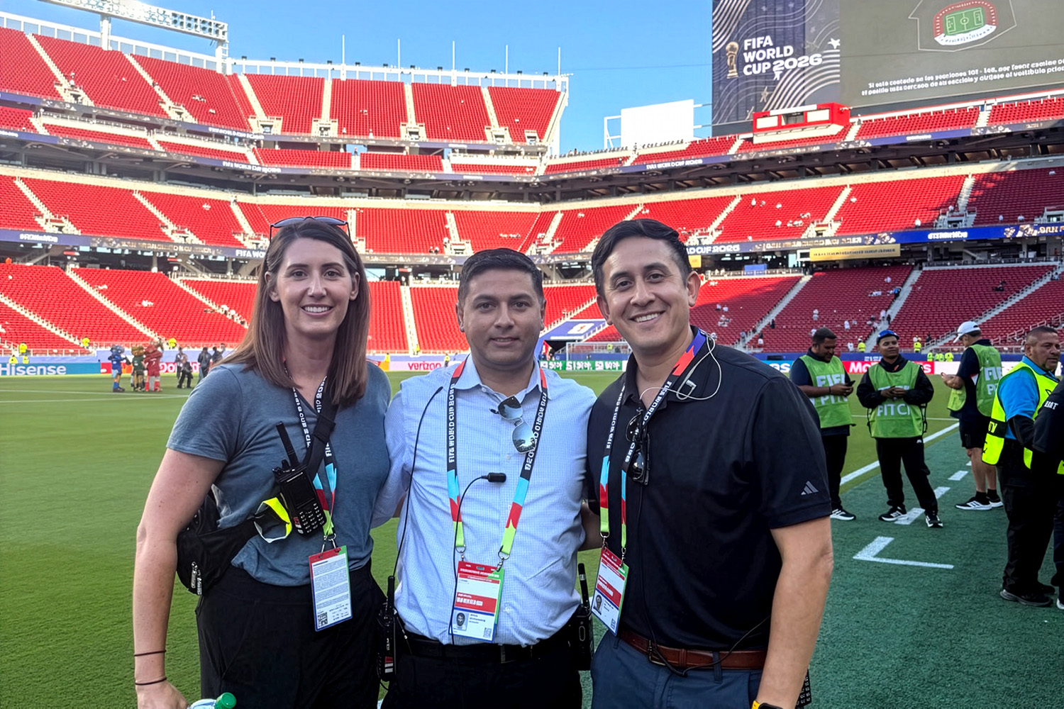 Alyssa Perry, Shashank Ravi, and Eli Carrillo smile together on the field of a stadium with empty red seats and World Cup signage.