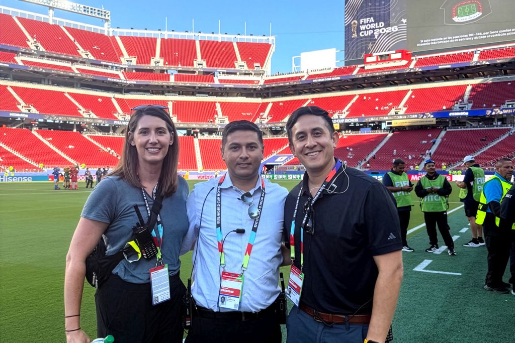 Alyssa Perry, Shashank Ravi, and Eli Carrillo smile together on the field of a stadium with empty red seats and World Cup signage.