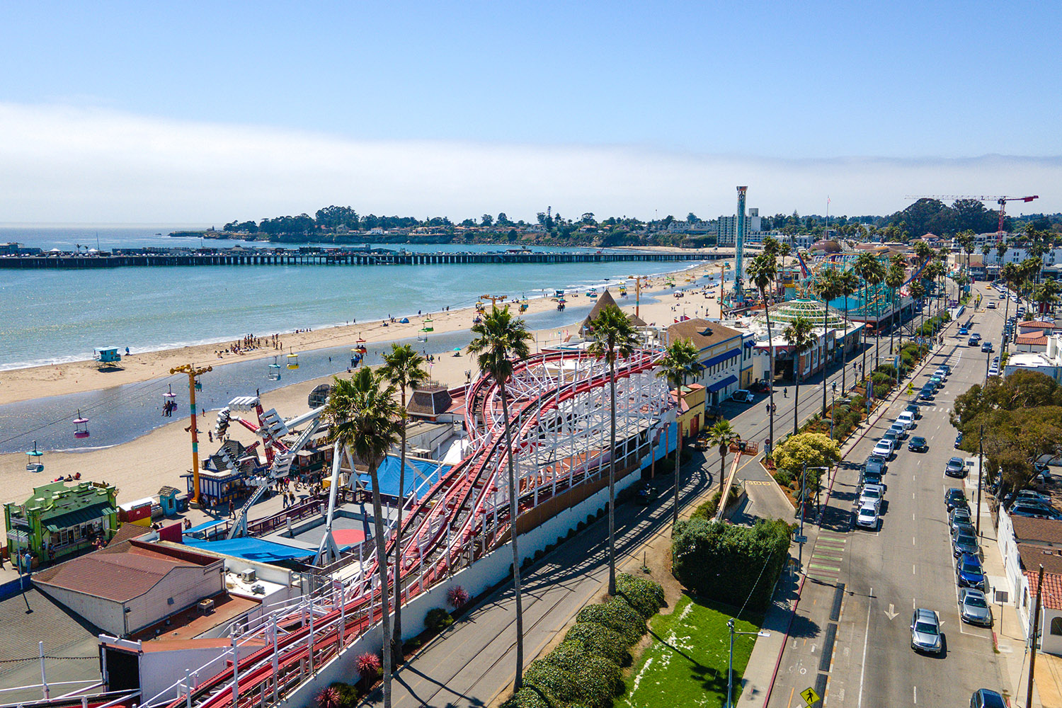 Aerial view of Santa Cruz boardwalk with a roller coaster and palm trees, alongside a sandy beach and ocean waves.