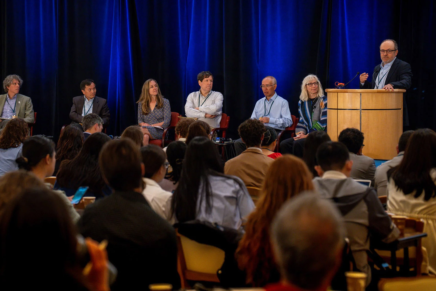 A panel of speakers engages an audience during a presentation, with one speaker at a podium discussing the topic.
