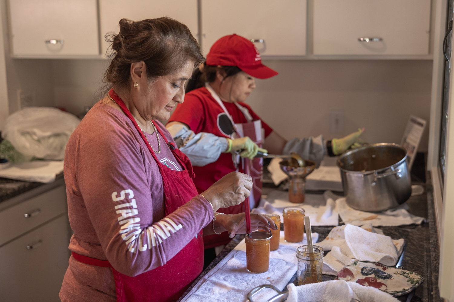 Two women in red aprons filling and sealing freshly made apricot jam into glass jars.