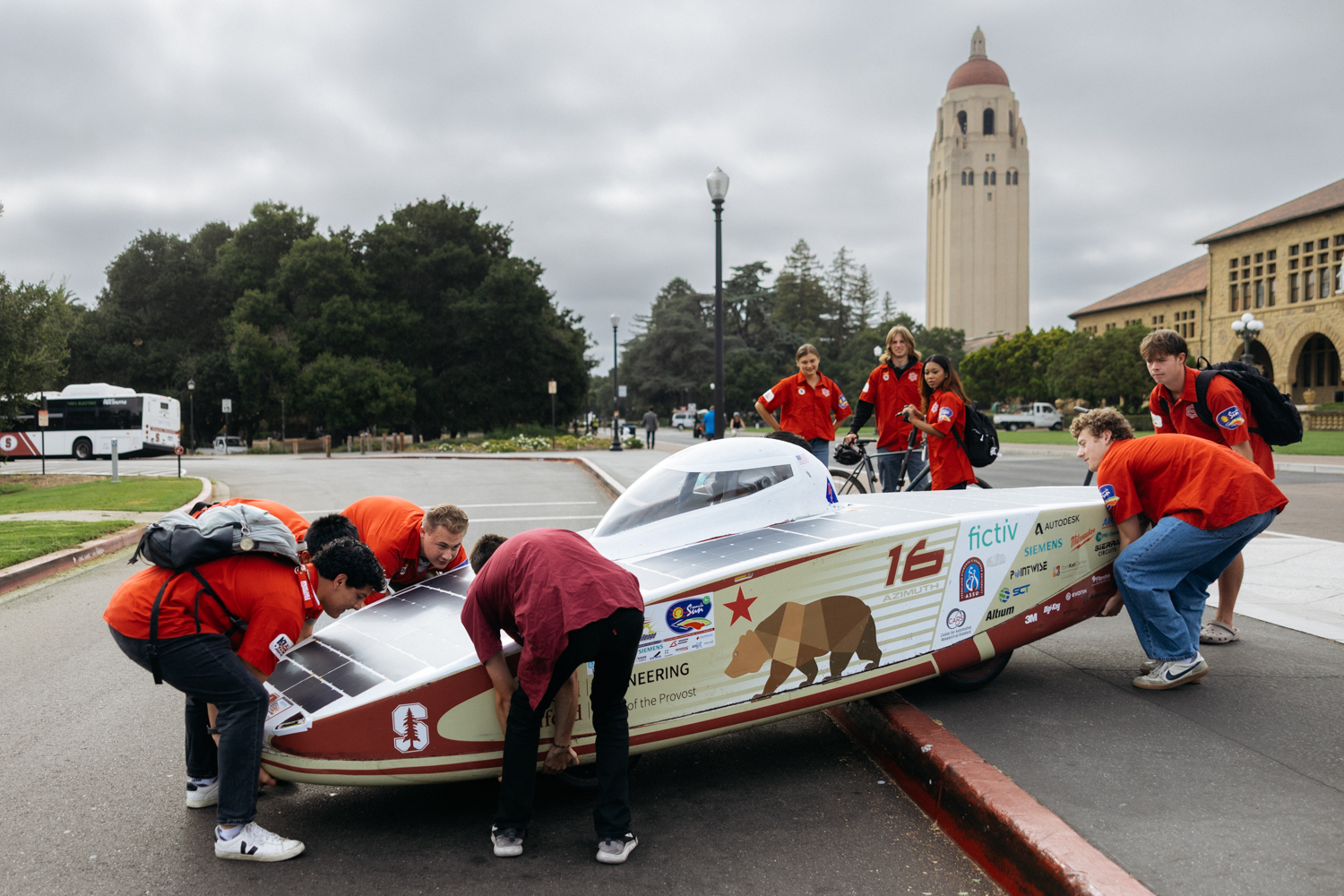 Stanford students in team shirts maneuver their solar car at the roadside, with Hoover Tower in the background.