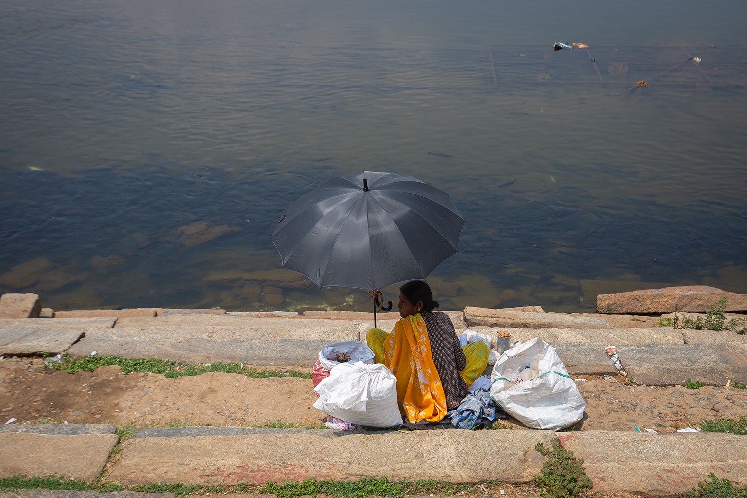 A woman sits by a riverbank under a black umbrella, surrounded by bags.