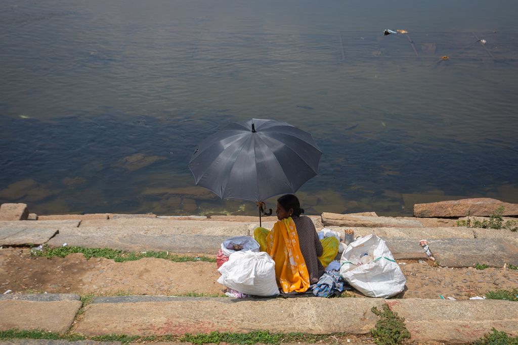 A woman sits by a riverbank under a black umbrella, surrounded by bags.