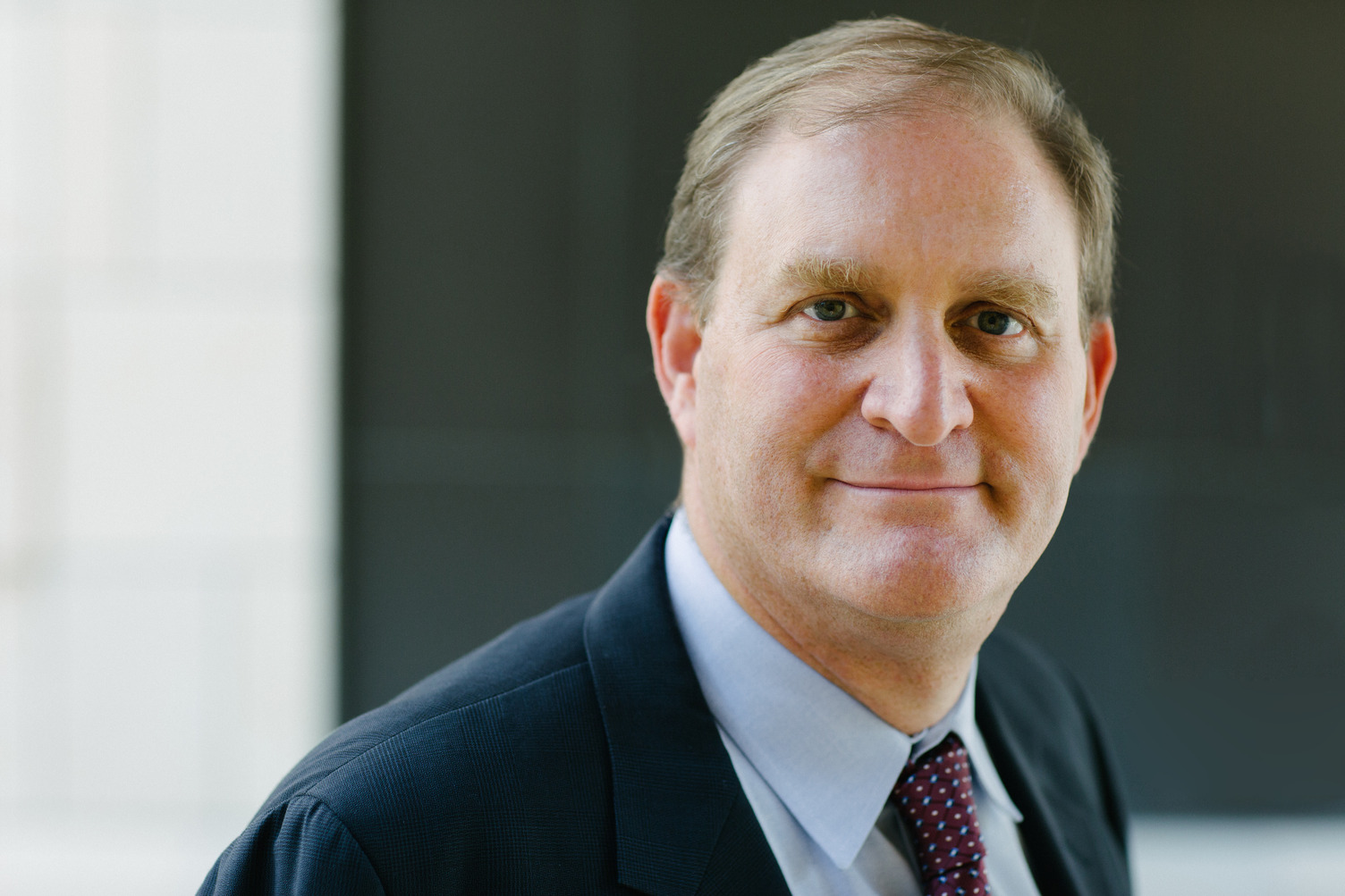 Nate Persily poses for a professional headshot, wearing a dark suit and tie against a blurred building backdrop.