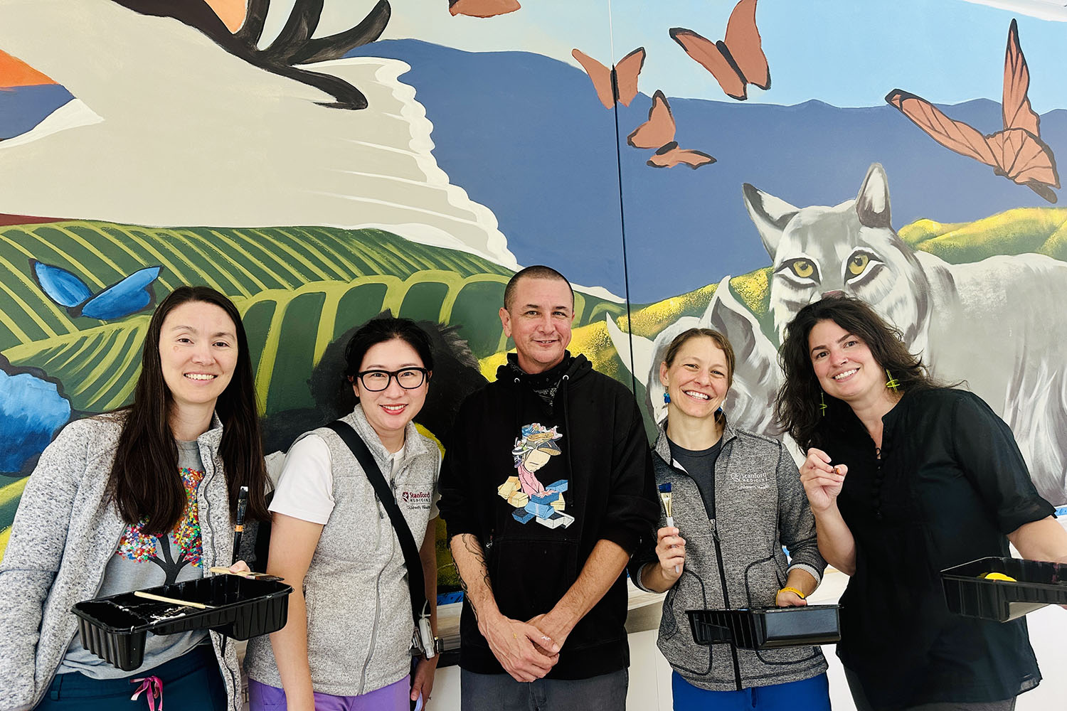 Five people pose with paint trays in front of a colorful wildlife mural showing a bobcat, butterflies, and rolling hills.