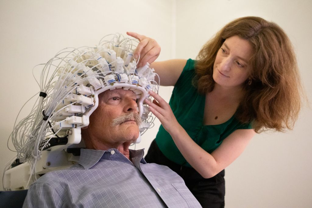 Open video A researcher fits a wired sensor cap used for brain imaging onto a seated study participant. in a modal