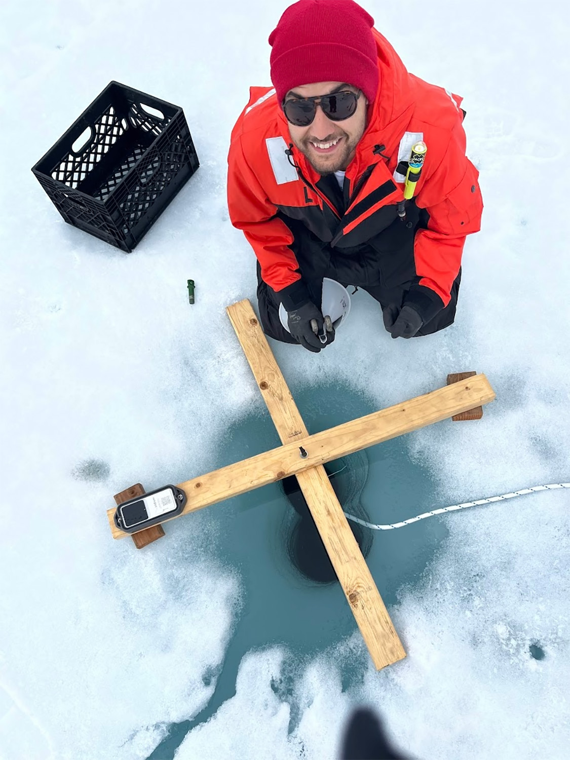 A researcher smiles while kneeling beside a hole cut in the ice for sampling equipment.