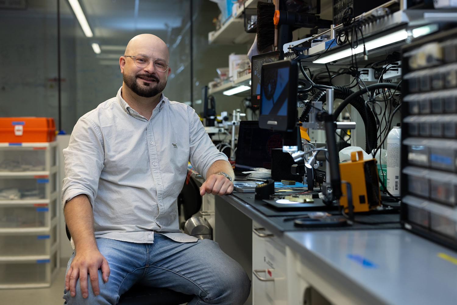 Mitchell Peterson sits beside a cluttered electronics lab bench with a digital microscope and tools.