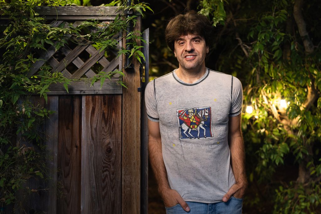 Open video Karl Deisseroth stands with his hands in his pockets beside a wooden garden gate at night, with string lights glowing behind him. in a modal