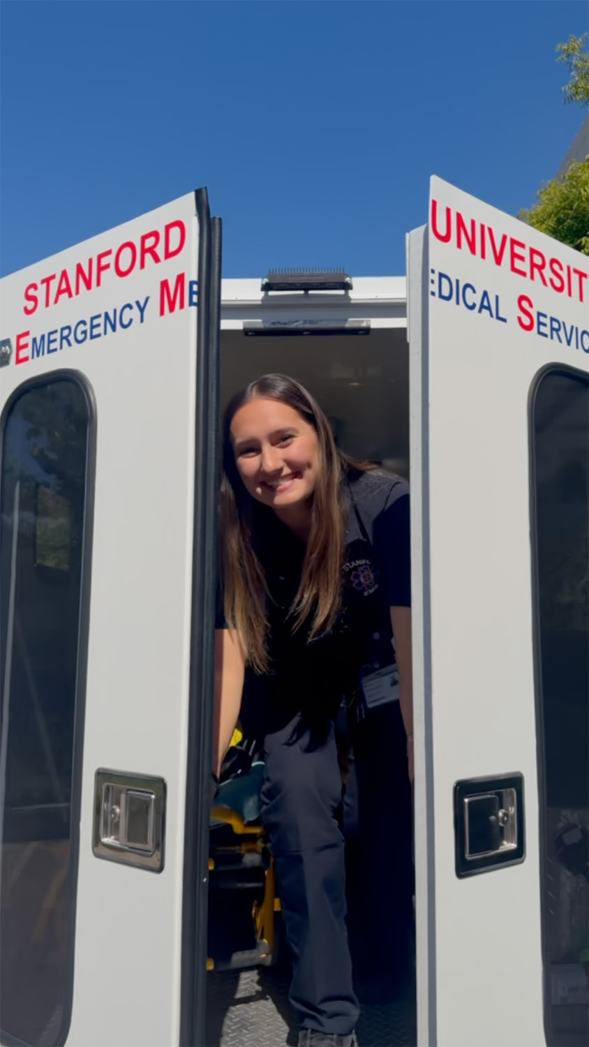 Smiling responder leans out from the open rear doors of an ambulance on a sunny day.