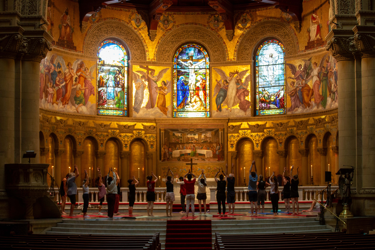 Participants raise their arms during an indoor yoga class at the front of Memorial church with golden mosaics and stained glass.