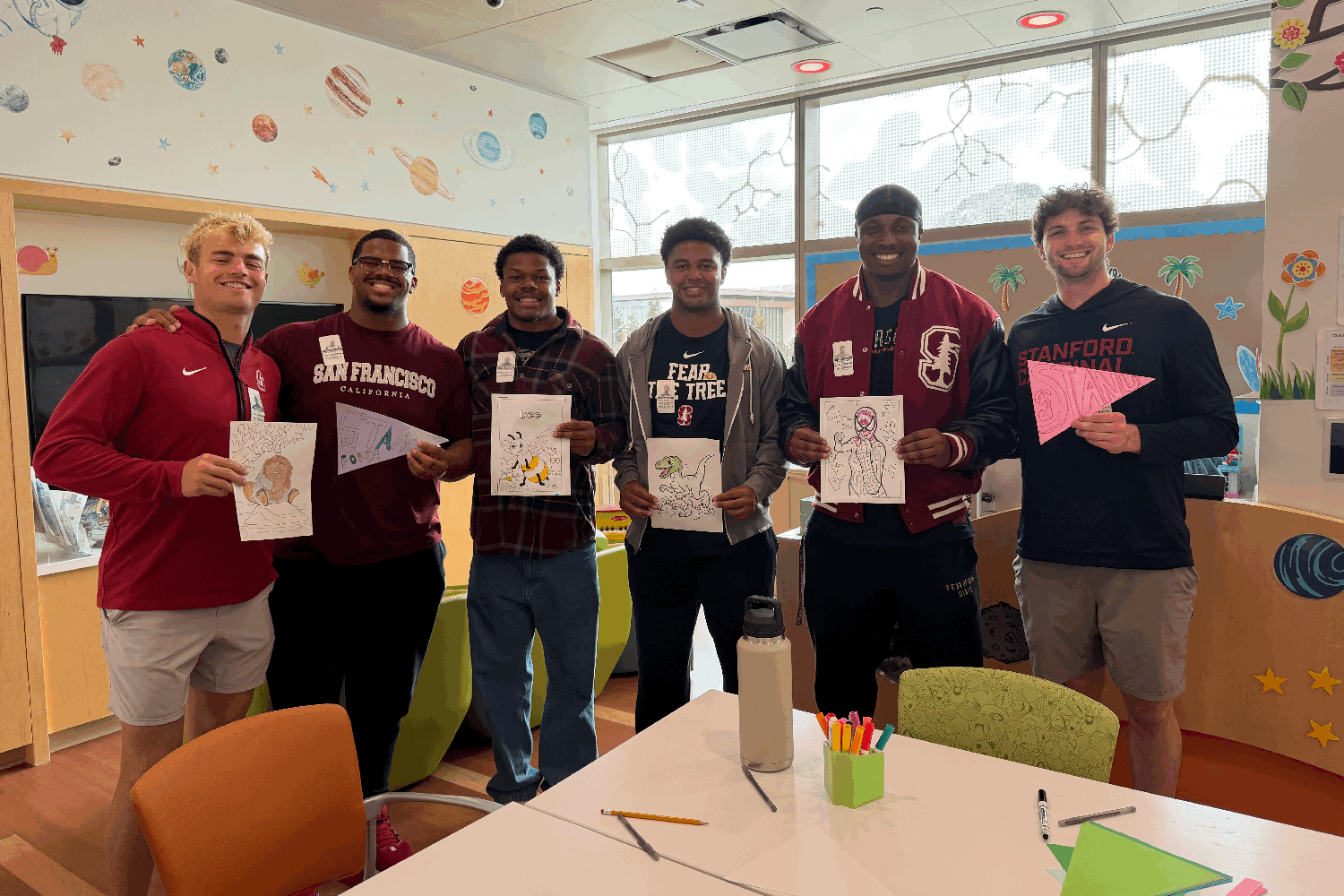 Six Stanford football players hold up coloring pages and paper pennants in a brightly decorated children’s hospital playroom.