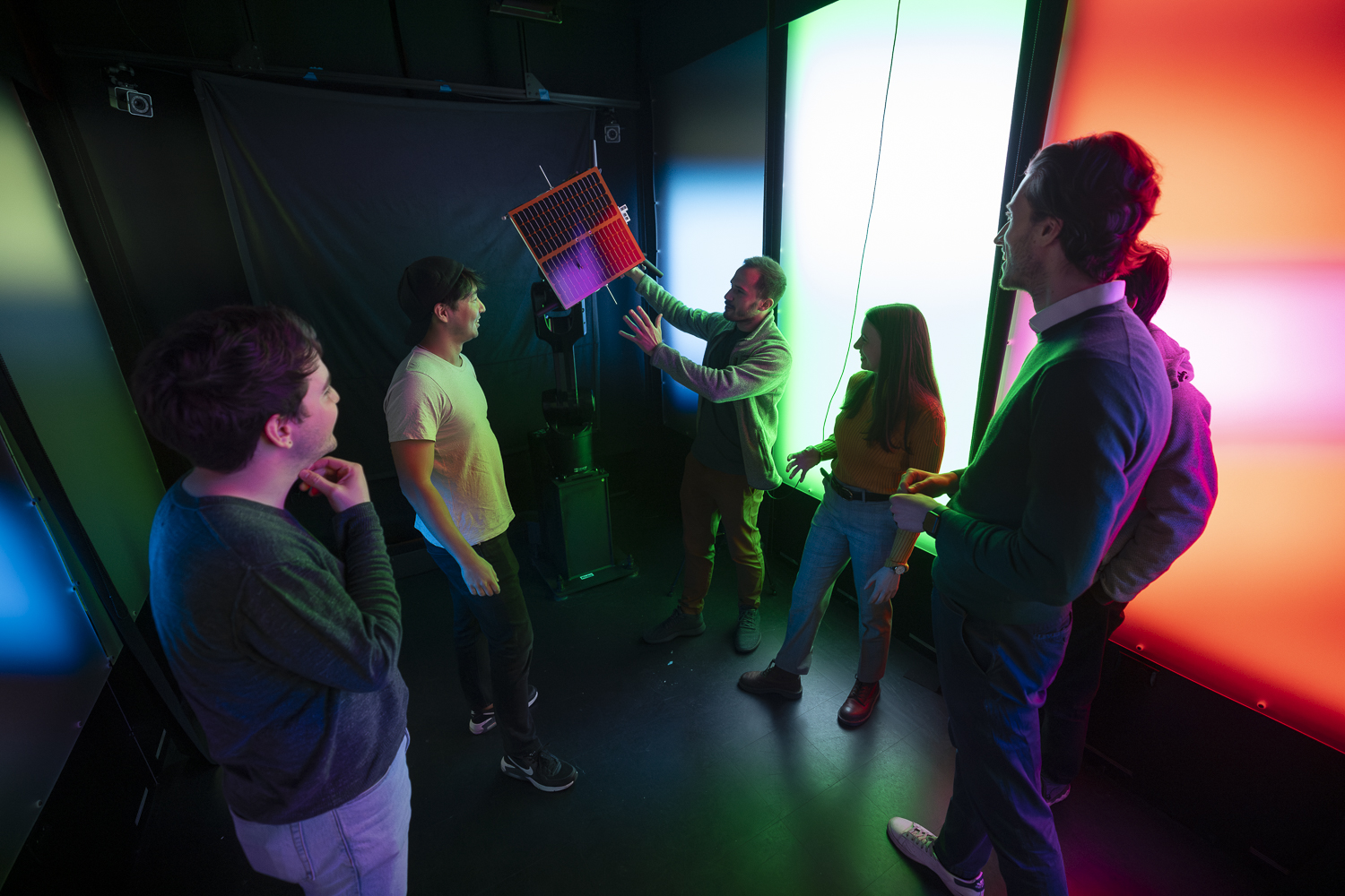 Six people stand inside a lab lit with multi-colored light panels on the walls. They are looking at soloar panel on a robotic arm