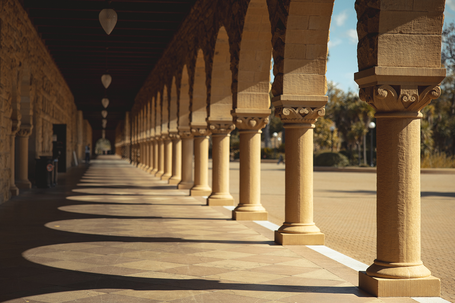 Carved sandstone columns cast geometric shadows upon a long Stanford arcade on a sunny day.