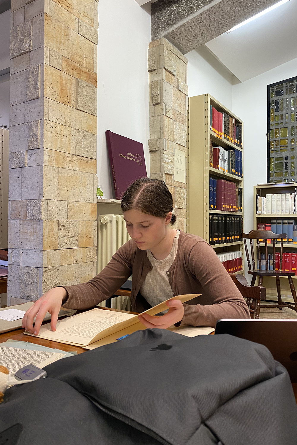 A student studies a book at a table in a library surrounded by shelves of books and stone pillars.