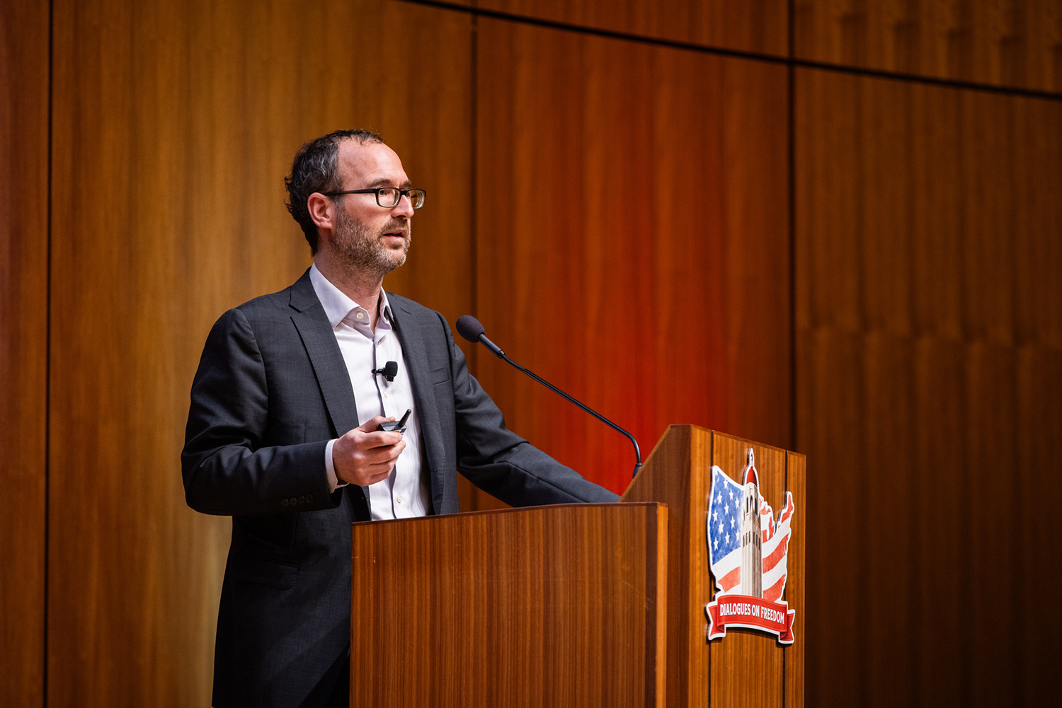 Dan Edelstein presenting at a podium, with a backdrop showing logos and American symbols.