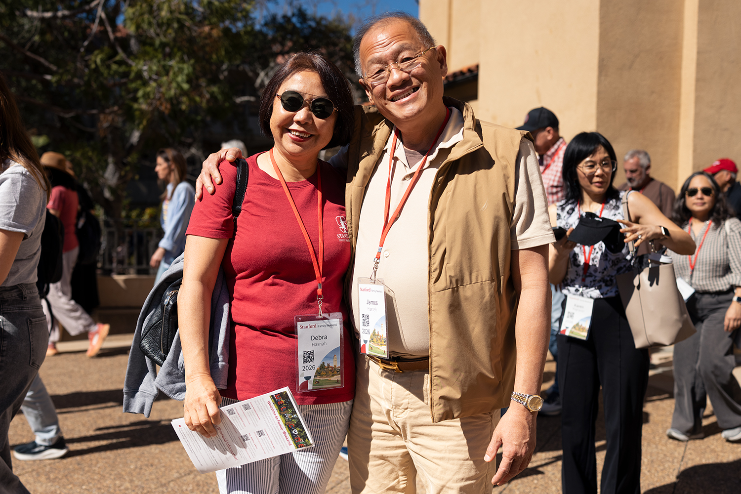 Two people pose for a photo during Family Weekend, wearing name badges; a group is visible in the background.