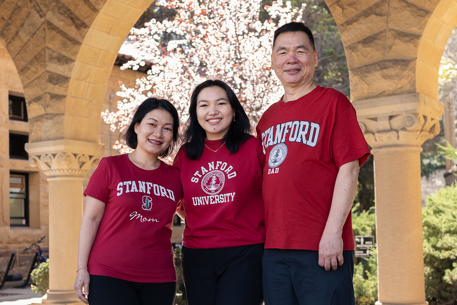 Emily Sidharta and her parents pose together outdoors, wearing Stanford-themed shirts, under an archway with blossoming trees in the background.