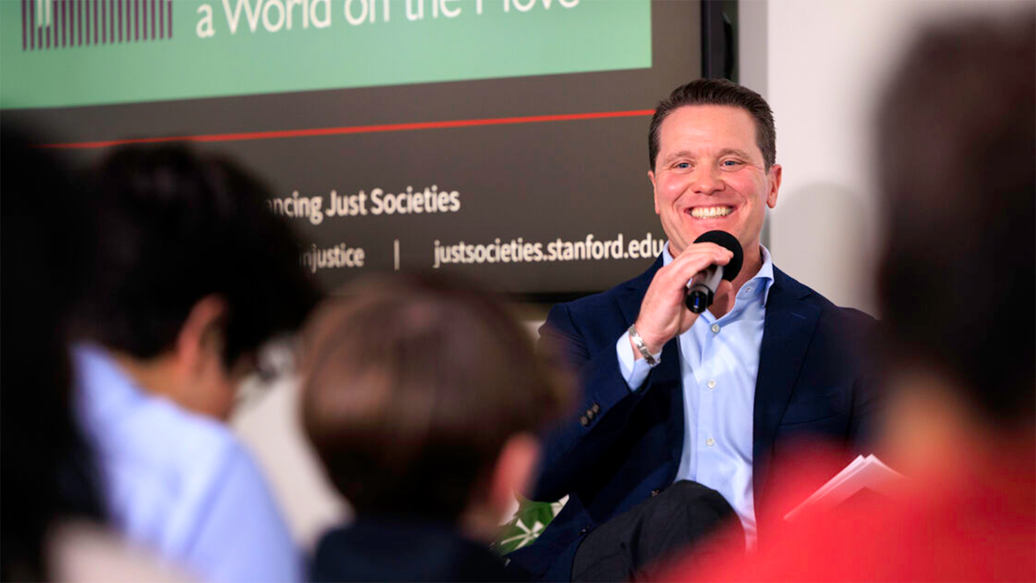 Tomás Jimenéz engages an audience during a discussion about advancing just societies, with a display board featuring relevant information in the background.