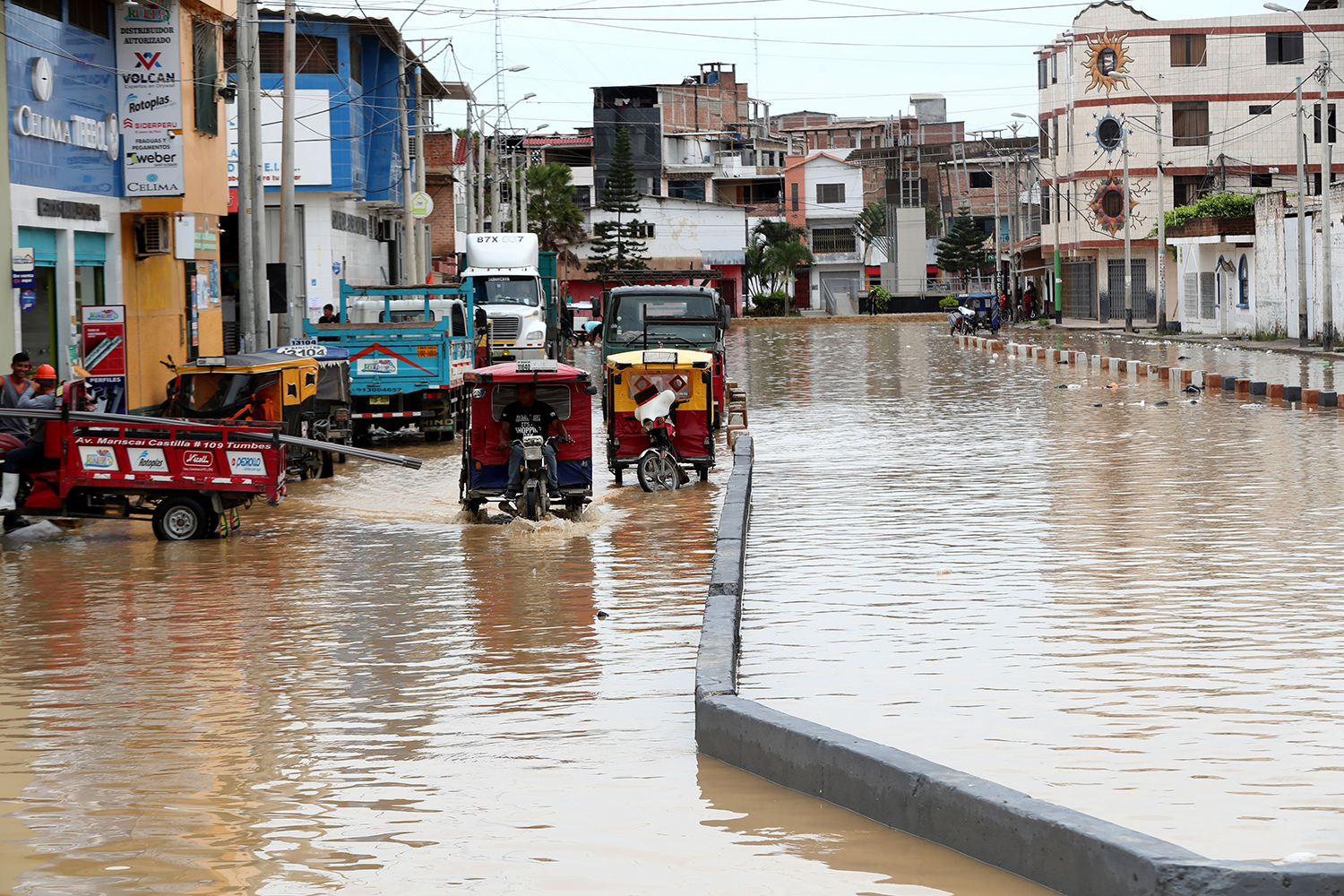 Vehicles navigate through a flooded street lined with buildings in an urban area affected by heavy rainfall.