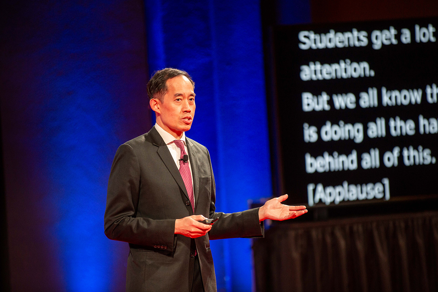 Joseph Woo in dark suit gestures while presenting on stage with text projected behind him.