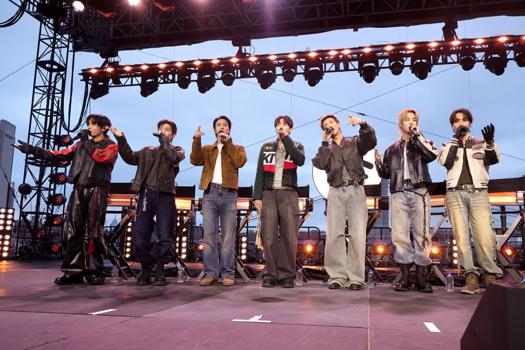 Seven performers of BTS stand in a line on an outdoor stage under bright lights during an evening concert.