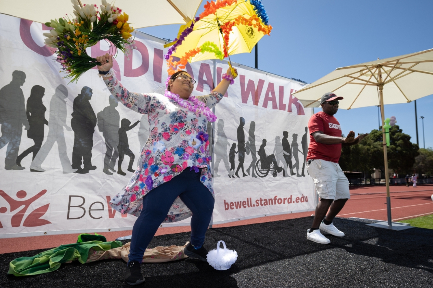 A person in a floral outfit dancing joyfully with an umbrella and flowers at a wellness event, with a banner in the background.