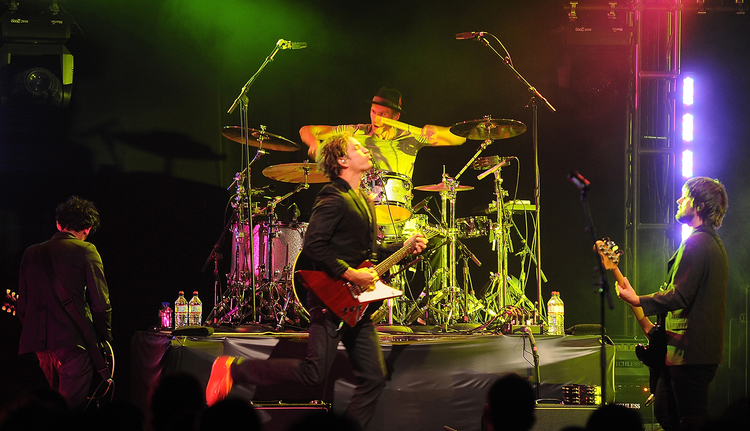 Third Eye Blind performing live on stage, with a guitarist, drummer, and other musicians illuminated by colorful lights.