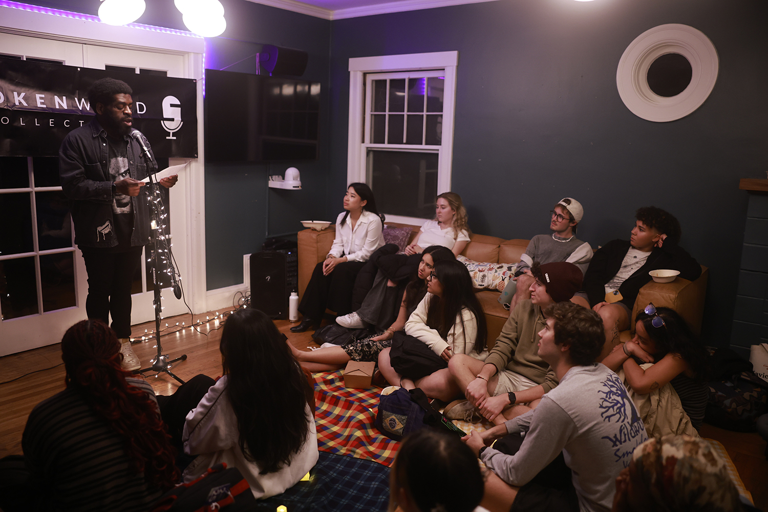 Hanif Abdurraqib standing in front of a microphone performing spoken word to a small room of people seated on couches and blankets.