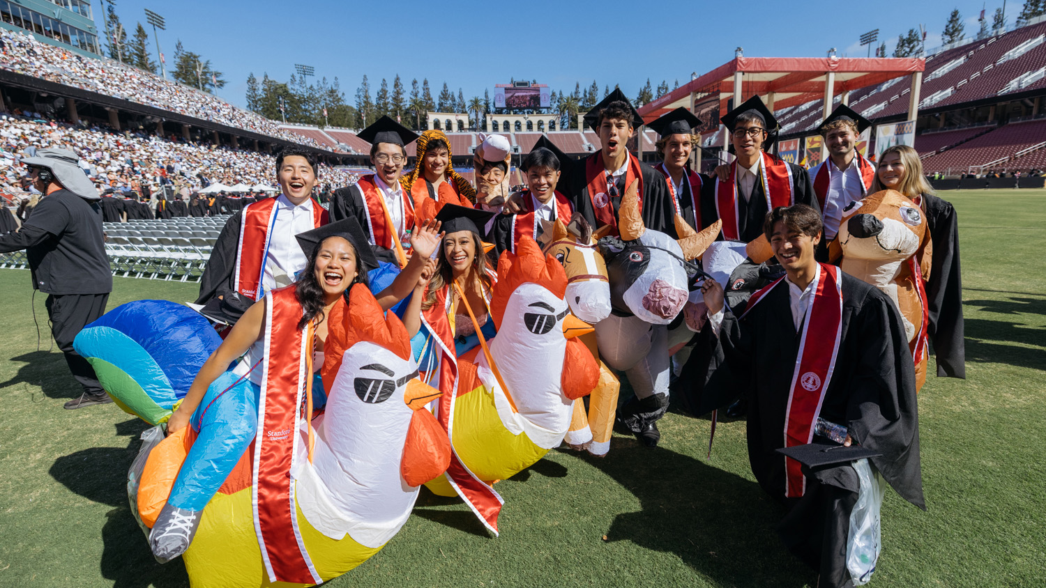 Graduates pose with inflatable animal costumes, celebrating at a graduation ceremony on a sunny day in a stadium.